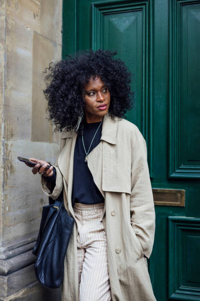 A stylish mid adult woman leaving her house in Newcastle upon Tyne, England. She is standing in front of her front door, using her phone while looking to her side with a contented look on her face as she heads off to work.